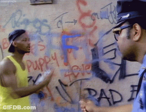 A Man In A Yellow Tank Top Talks To A Police Officer In Front Of A Wall Covered In Graffiti Including The Word Puppy GIF