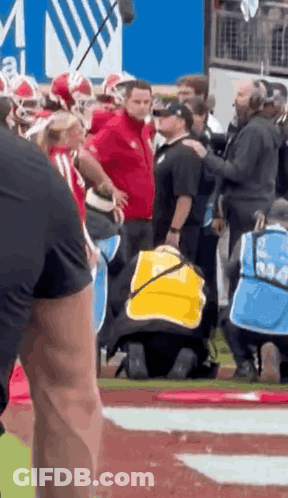 A Man In A Yellow Vest Is Kneeling On A Football Field With A Group Of People Standing Around Him . GIF