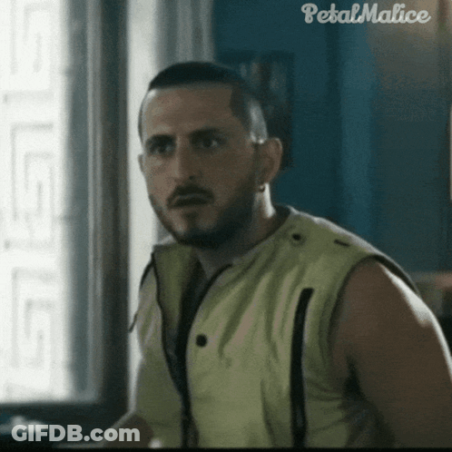 A Man In A Yellow Vest Is Standing In Front Of A Window With Petal Malice Written On The Bottom Right Corner GIF
