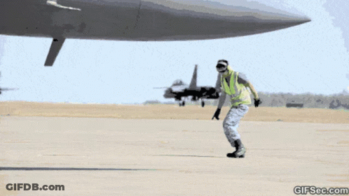 A Man In A Yellow Vest Is Standing In Front Of An Airplane With A Gifsec.com Watermark GIF