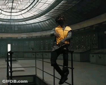 A Man In A Yellow Vest Sits On A Railing In An Empty Room GIF