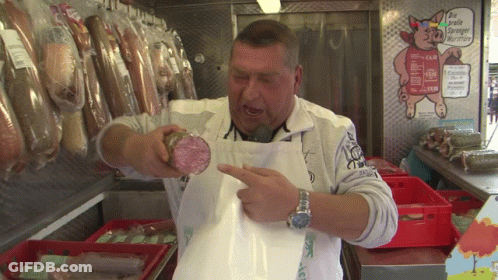 A Man In An Apron Holds A Piece Of Meat In Front Of A Sign That Says " Die Größte Europäische Wurst " GIF