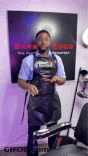 A Man In An Apron Is Standing In Front Of A Barber Chair Holding A Comb . GIF