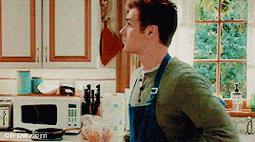 A Man In An Apron Stands In A Kitchen GIF