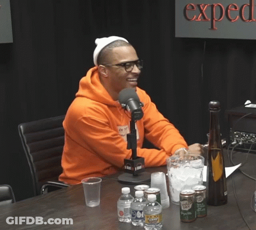 A Man In An Orange Hoodie Sits In Front Of A Microphone In Front Of A Sign That Says " Expect " GIF