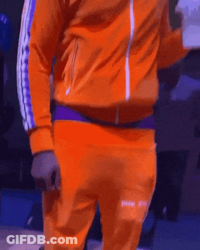 A Man In An Orange Jacket And Orange Sweatpants Is Standing On A Stage GIF
