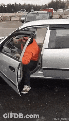A Man In An Orange Jacket Is Getting Out Of A Silver Car With The Door Open GIF
