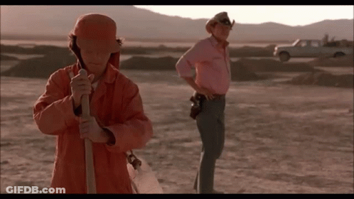 A Man In An Orange Jacket Is Holding A Shovel In The Desert While Another Man In A Pink Shirt Stands Behind Him . GIF