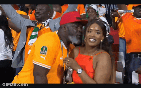 A Man In An Orange Puma Shirt Stands Next To A Woman In An Orange Top GIF