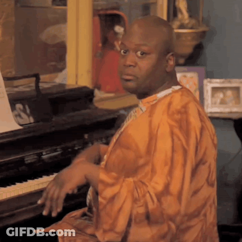 A Man In An Orange Robe Sits At A Piano With A Sign That Says S On It GIF