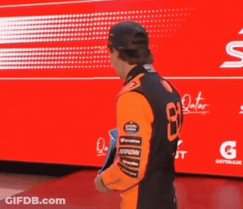 A Man In An Orange Shirt And Hat Is Holding A Trophy In Front Of A Red Background . GIF