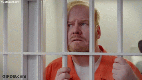 A Man In An Orange Shirt Is Behind Bars In A Prison Cell . GIF