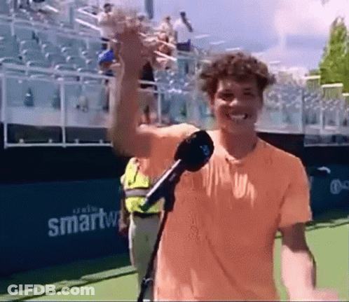 A Man In An Orange Shirt Is Holding A Microphone On A Tennis Court GIF