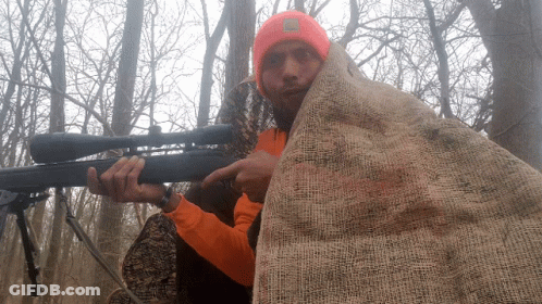 A Man In An Orange Shirt Is Holding A Rifle And Pointing GIF