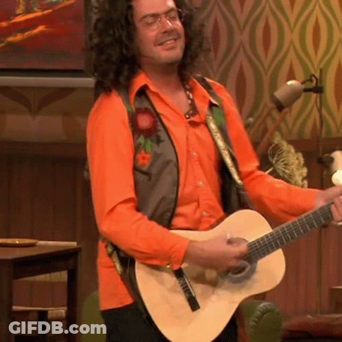 A Man In An Orange Shirt Is Playing A Guitar With A Strap That Says ' Fender ' On It GIF