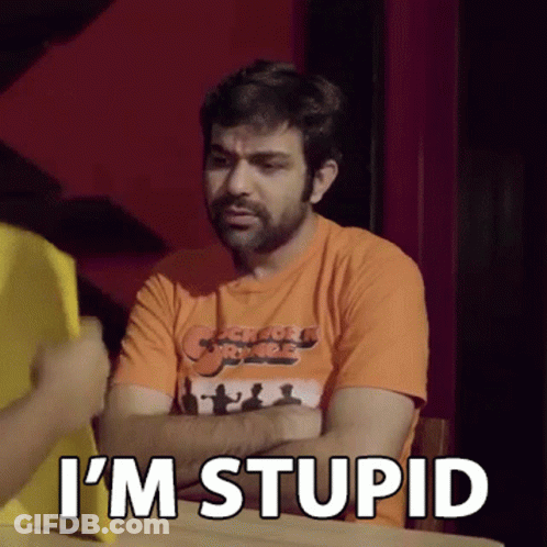A Man In An Orange Shirt Is Sitting At A Table With His Arms Crossed And Says `` I 'M Stupid '' GIF