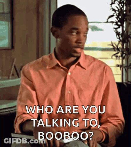 A Man In An Orange Shirt Is Sitting In Front Of A Window And Says Who Are You Talking To Booboo ? GIF