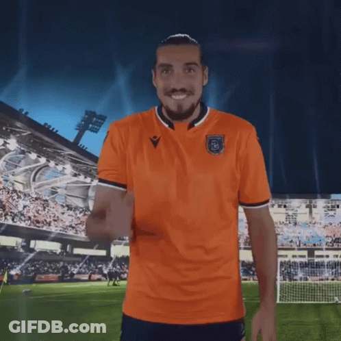 A Man In An Orange Shirt Is Standing On A Soccer Field With His Arms Outstretched GIF