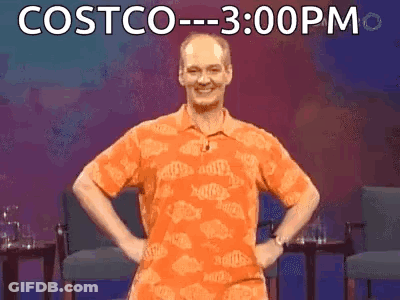 A Man In An Orange Shirt Is Standing On A Stage With His Hands On His Hips And The Words Costco 3:00 Pm Behind Him GIF
