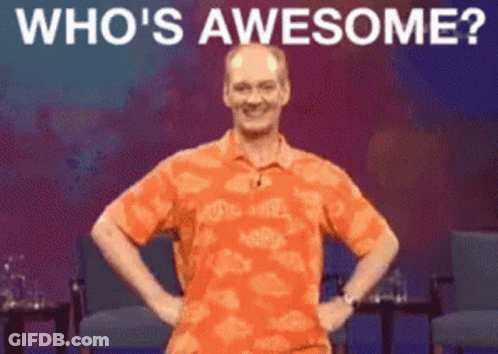 A Man In An Orange Shirt Is Standing On A Stage With His Hands On His Hips And The Words " Who 'S Awesome " Above Him GIF