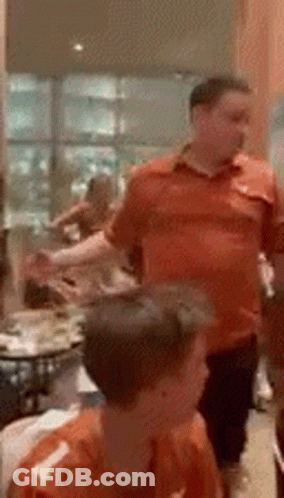 A Man In An Orange Shirt Stands In A Room With Other People GIF
