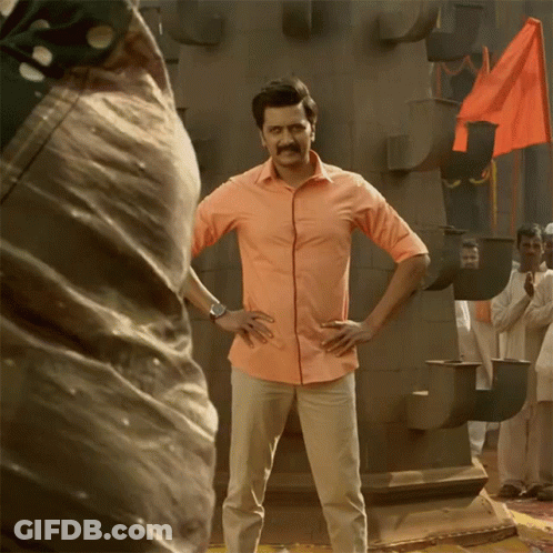 A Man In An Orange Shirt Stands With His Hands On His Hips In Front Of A Crowd GIF