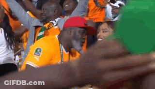 A Man In An Orange Shirt With The Letter C On It Is Sitting In A Crowd Of People . GIF