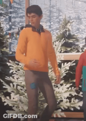 A Man In An Orange Sweater And Brown Pants Is Dancing In Front Of A Painting Of Snow Covered Trees . GIF