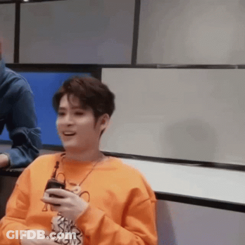A Man In An Orange Sweater Is Holding A Cell Phone In His Hand And Smiling . GIF