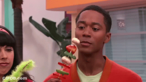A Man In An Orange Sweater Is Holding A Skewer Of Vegetables In His Mouth GIF