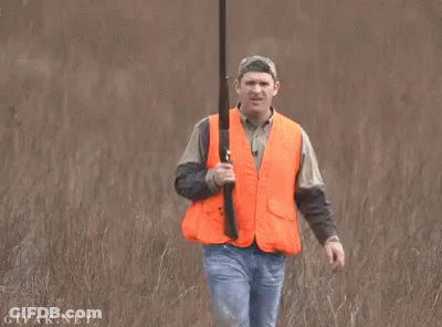 A Man In An Orange Vest Is Holding A Shotgun In A Field GIF