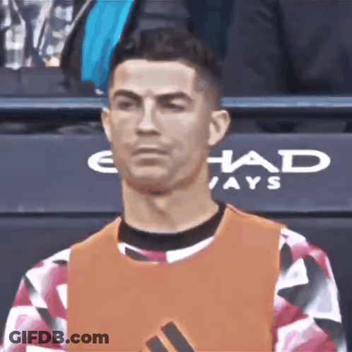 A Man In An Orange Vest Is Sitting In A Stadium And Looking At The Camera . GIF