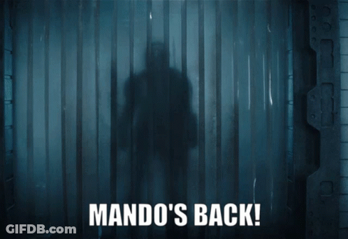 A Man In Armor Is Standing In A Dark Room With The Words Mando 'S Back On The Bottom GIF