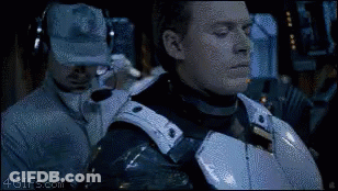A Man In Armor Is Standing Next To A Man Wearing A Hat And Headphones . GIF
