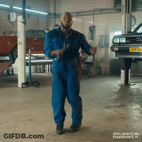 A Man In Blue Overalls Is Dancing In A Garage GIF