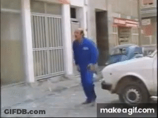 A Man In Blue Overalls Is Walking Down The Street Next To A White Car GIF