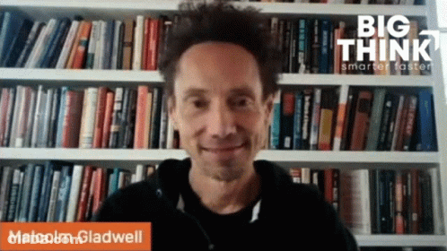 A Man In Front Of A Bookshelf With The Name Malcolm Gladwell GIF