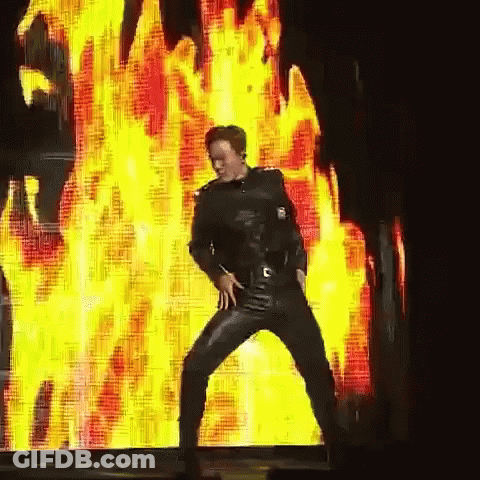 A Man In Leather Pants Is Dancing In Front Of A Large Screen With Flames On It GIF