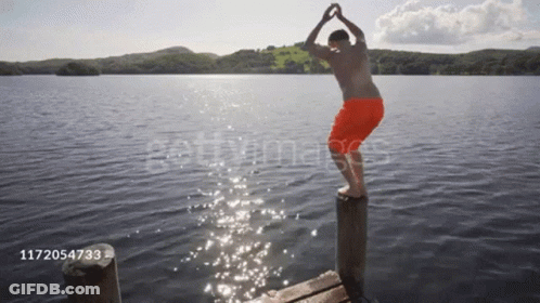 A Man In Orange Shorts Is Jumping Into A Lake From A Pier GIF