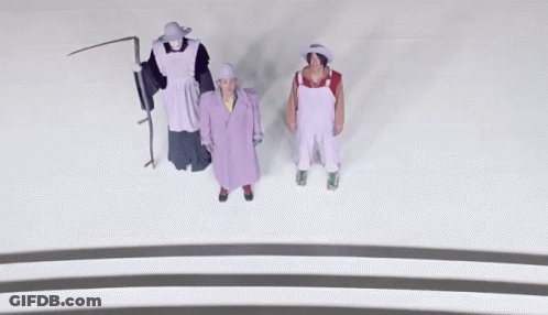A Man In Overalls And A Woman In A Purple Coat Are Standing Next To Each Other GIF