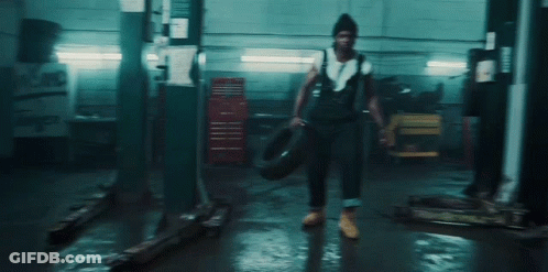 A Man In Overalls Is Holding A Tire In A Dark Garage . GIF