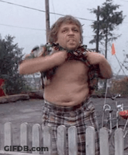 A Man In Plaid Shorts Is Standing In Front Of A Fence And Taking Off His Shirt GIF
