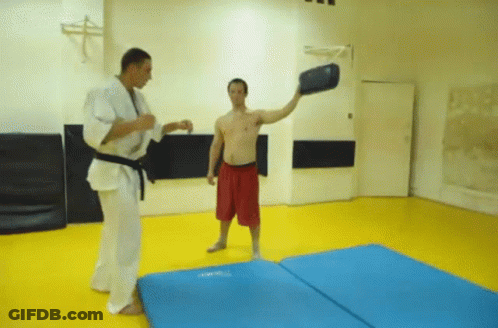A Man In Red Shorts Is Standing On A Blue Mat While Another Man Throws A Black Object GIF