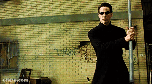 A Man In Sunglasses Holds A Pole In Front Of A Brick Wall With Graffiti On It That Says Dman GIF