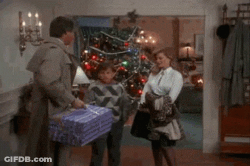 A Man Is Carrying A Purple Gift Box In Front Of A Christmas Tree GIF