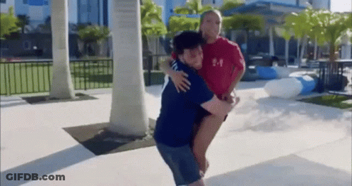 A Man Is Carrying A Woman On His Back In A Park While A Woman Looks On . GIF