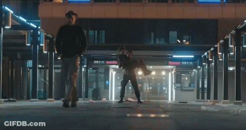 A Man Is Carrying Another Man In His Arms In A Building With A Sign That Says Donuts GIF