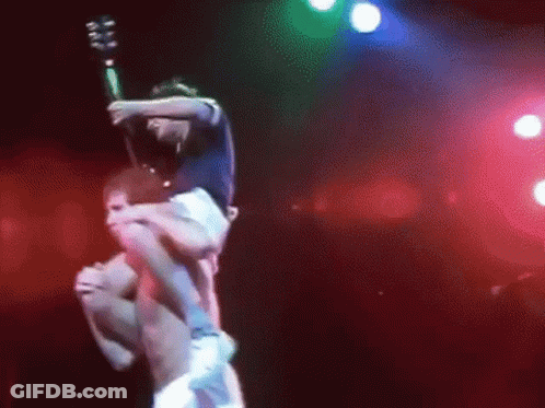 A Man Is Carrying Another Man On His Shoulders On A Stage While Holding A Guitar . GIF