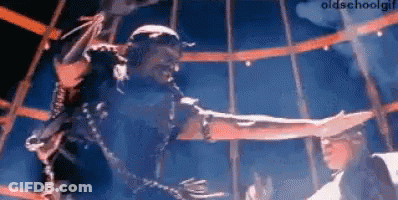 A Man Is Chained To A Pole And Reaching Out Towards Another Man . GIF