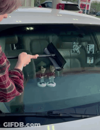 A Man Is Cleaning The Windshield Of A Car With A Wiper And Two Bobbleheads On It GIF